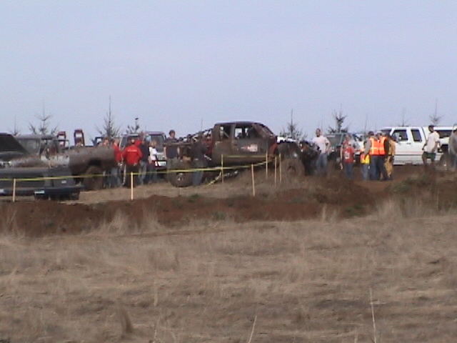 Mud-covered lifted pickup behind yellow caution tape with spectators at an off-road event