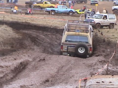 1994 Ford Explorer - An off-road event features a tan SUV struggling in a muddy trench, with other vehicles and spectators in the background.