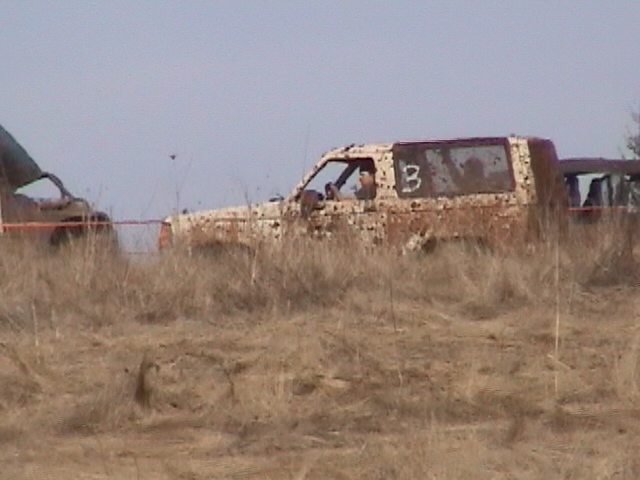 Mud-spattered SUV marked with a 3 sitting in tall dry grass at an event