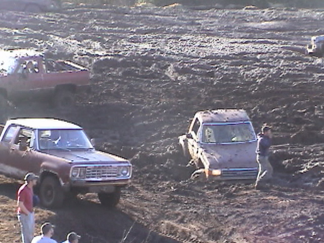 White SUV stuck in a churned mud pit with pickups and spectators nearby