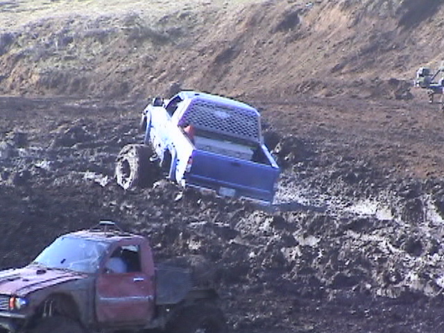Blue and white lifted mud truck climbing out of a mud pit, red mud truck in foreground