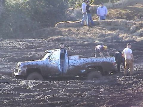 1994 Ford Explorer - A pickup truck is stuck deep in mud at a festival, with several people attempting to help.