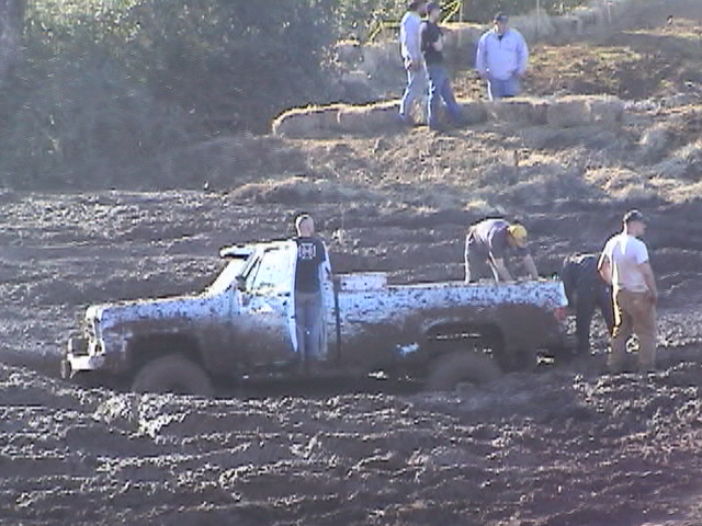 White square-body pickup stuck in a mud pit with spectators standing around it