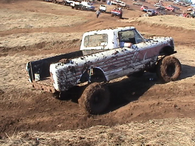 Mud-covered pickup truck with large tires on a dirt track, other vehicles in background
