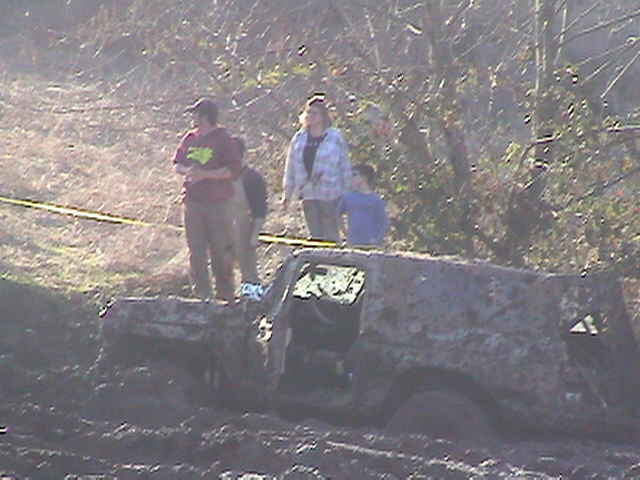 Mud-covered SUV stuck door-open in a deep mud pit with spectators watching