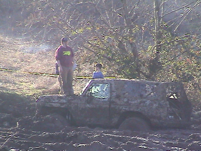 Topless mud-covered 4x4 stuck to its rockers in a mud pit with spectators behind