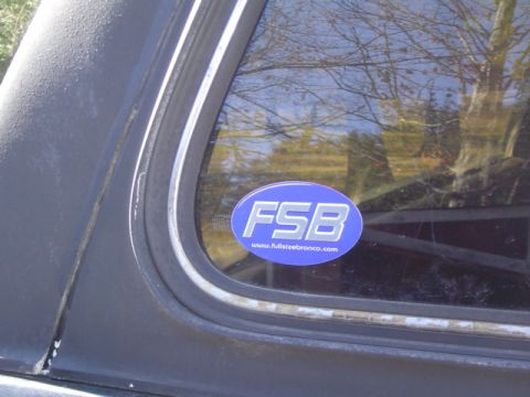 1979 Ford Bronco - Close-up of a blue oval "FSB: www.fullsizebronco.com" decal applied to the inside of a tinted quarter window on a black vehicle. Weathered…