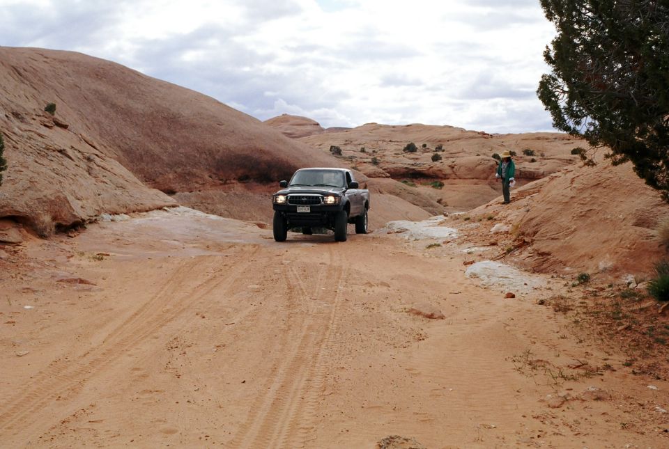 Dark pickup crossing a sandy desert trail among red rock with a hiker watching