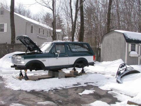 1994 Ford Bronco - A two-tone green and white Ford Bronco is elevated on jack stands in a snowy driveway, with its hood open.