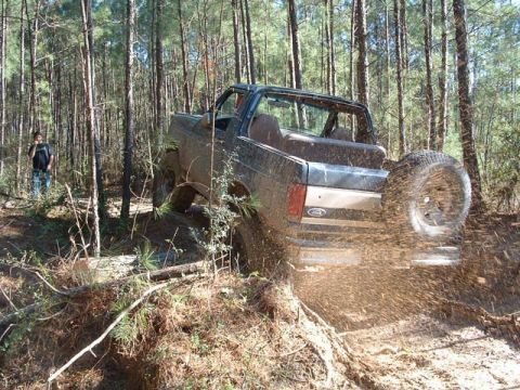 1993 Ford Bronco - A Ford Bronco navigates a muddy trail through a dense forest, kicking up dirt as it goes.