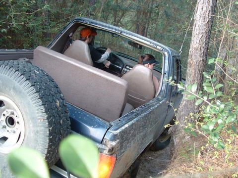 1993 Ford Bronco - A dark blue 1993 Ford Bronco navigates a wooded trail, showing its interior and spare tire.