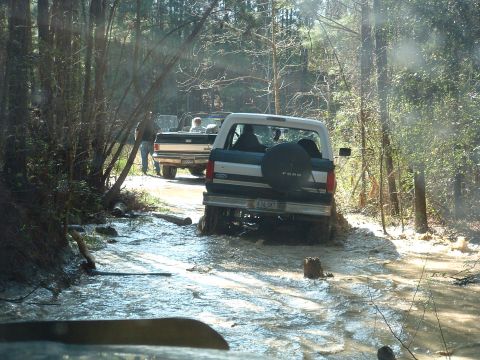 1993 Ford Bronco - Two Ford vehicles navigate a challenging off-road course, splashing through mud and water.