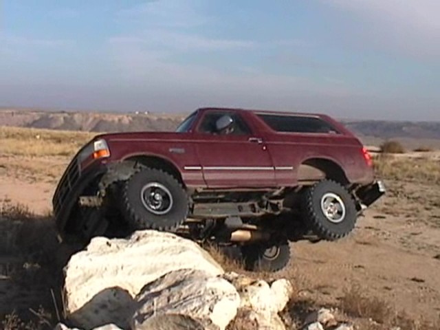 Maroon lifted Ford Bronco crawling nose-down over large white rocks in a desert area