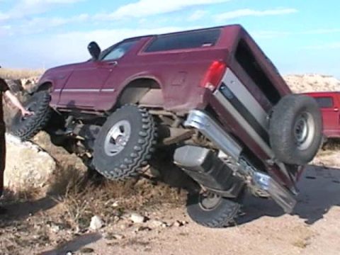 1993 Ford Bronco - This maroon Ford Bronco is tilted on its side, with its large off-road tires struggling for traction on rocky terrain.