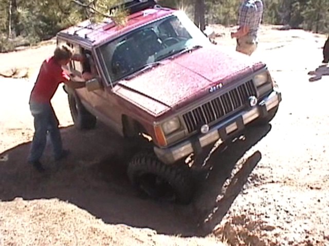 Person in red shirt spotting a dark red lifted Jeep Cherokee through a muddy rut