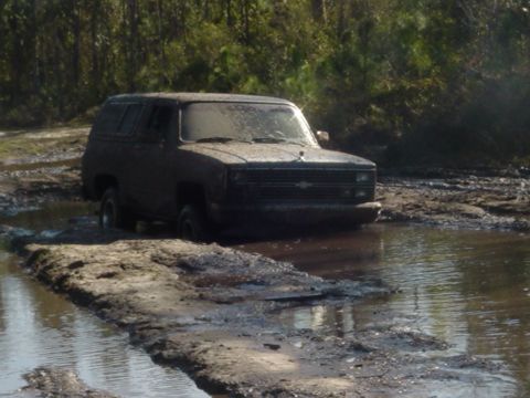 1989 Ford Bronco - A dark-colored SUV navigates through a muddy water obstacle, its front grille partially submerged.