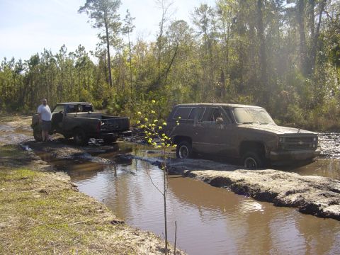 1989 Ford Bronco - Two off-road vehicles are stuck in a muddy water crossing in a wooded area.