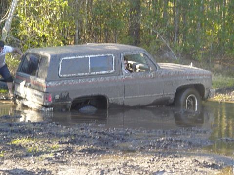 1989 Ford Bronco - This SUV is stuck deep in the mud and water, with a person attempting to help from the rear.