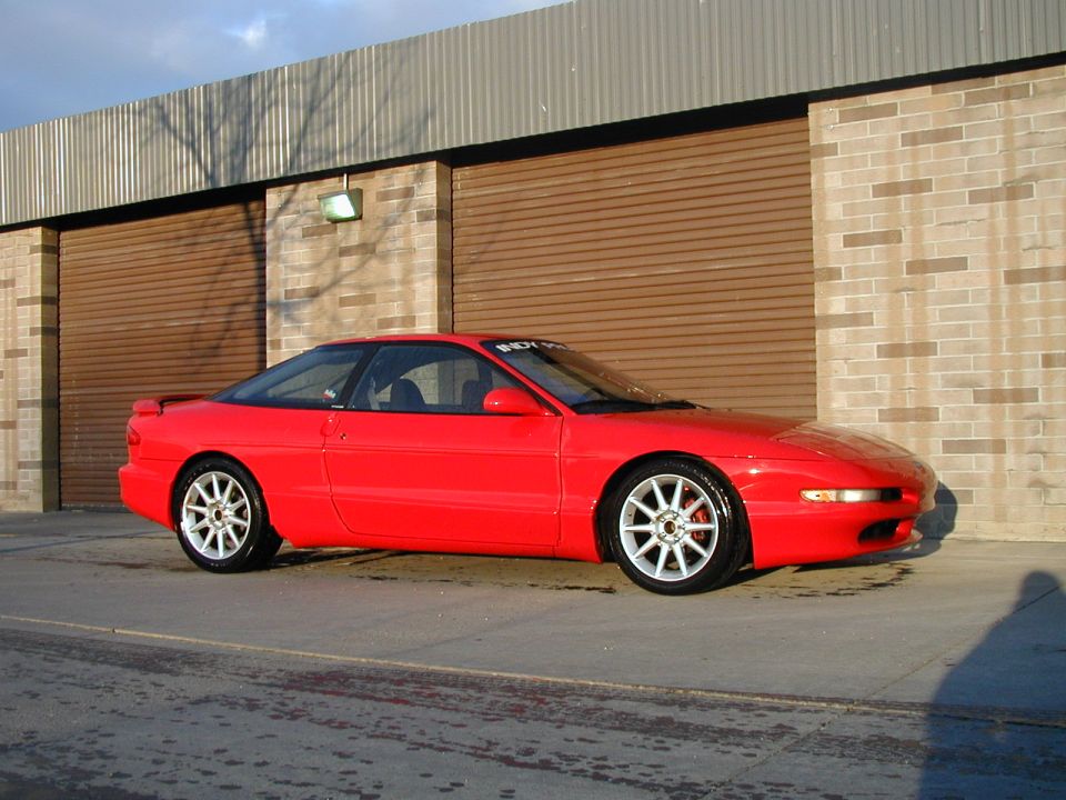 Red Ford Probe coupe with white wheels parked in front of brick building with roller doors