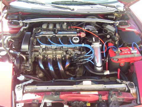 1997 Ford Probe - The engine bay of a red car shows a V6 engine with aftermarket modifications and a red Optima battery.