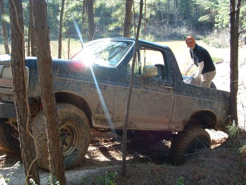 1993 Ford Bronco - A person in an orange hat is in the bed of a muddy Ford Bronco navigating a wooded area.