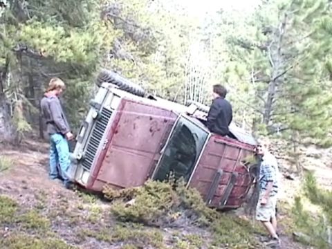 1993 Ford Bronco - A red SUV lies on its side in a forest clearing, with three individuals observing the scene.