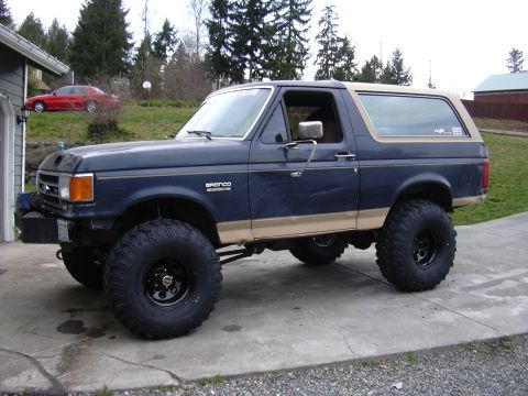 1991 Ford Bronco - Dark blue Ford Bronco with tan lower two-tone and tan hardtop, photographed from the front three-quarter on a concrete driveway with evergr…