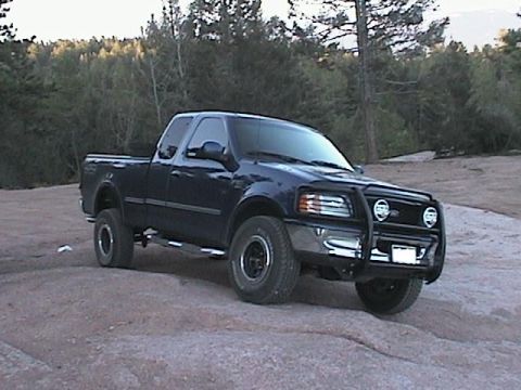 1993 Ford Bronco - This dark blue pickup truck, equipped with a prominent bull bar, is parked on a rocky surface in a wooded area.