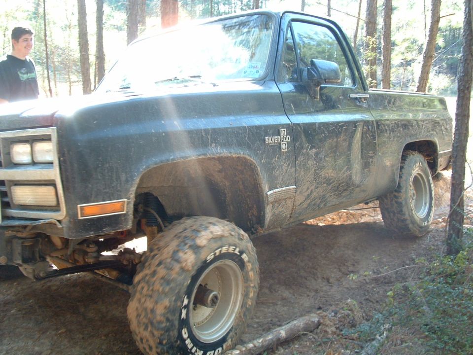 Muddy, lifted 1981 Chevrolet Silverado pickup truck on a wooded trail. A person stands near the front fender
