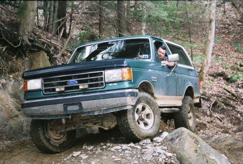 1990 Ford Bronco - Driver steers a green 1990 Ford Bronco through a challenging off-road path in a forest.