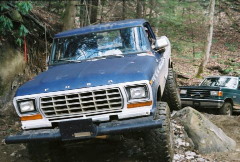 1990 Ford Bronco - Two trucks navigate a challenging off-road course through a wooded area.