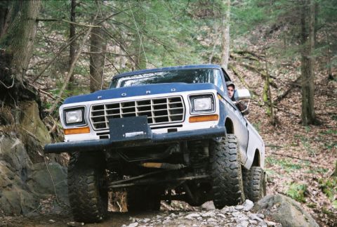 1990 Ford Bronco - A blue and white Ford Bronco navigates a challenging off-road path through a wooded area.