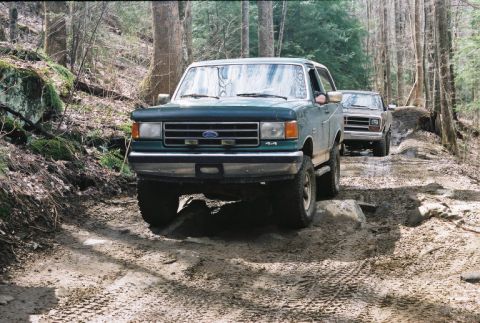 1990 Ford Bronco - Two Ford trucks navigate a rocky, muddy path through a wooded area.