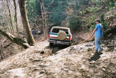 1990 Ford Bronco - A man walks beside a Ford Bronco navigating a challenging off-road path in a forest.