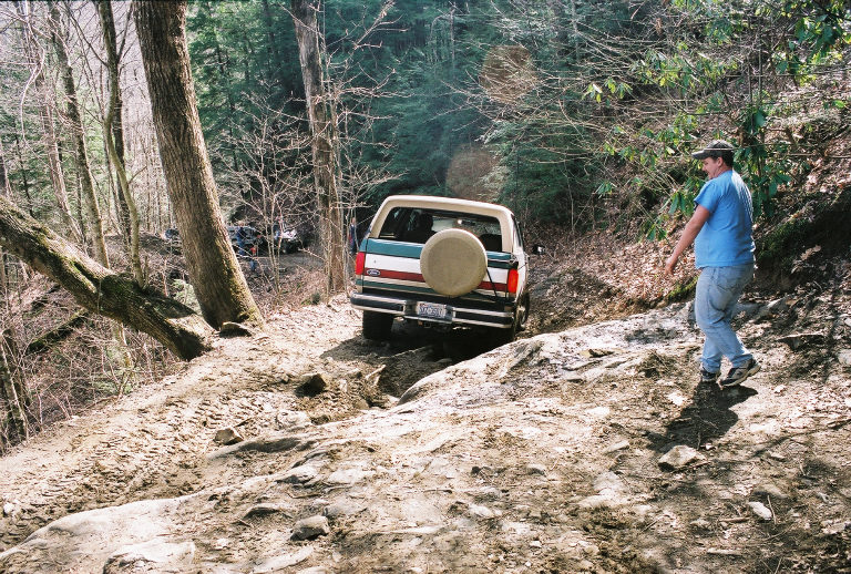 Tan and teal Ford Bronco with a spare tire on the back, driving on a rocky, wooded trail