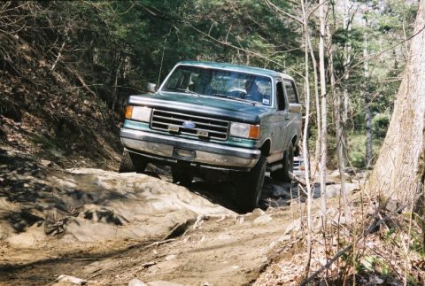 1990 Ford Bronco - A Ford Bronco navigates a rocky off-road trail surrounded by trees.
