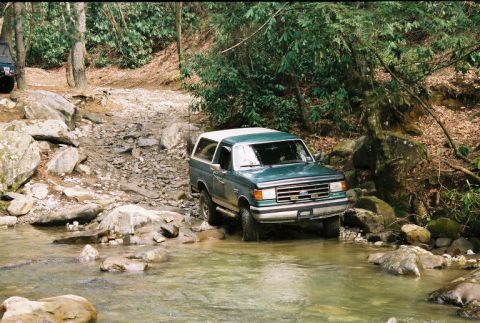 1990 Ford Bronco - This Ford Bronco navigates a shallow creek bed surrounded by trees and large rocks.
