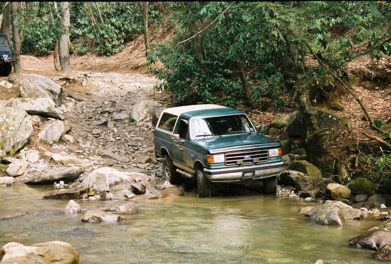 Green Ford Bronco crossing a rocky stream in a wooded area. Another vehicle is partially visible to the left