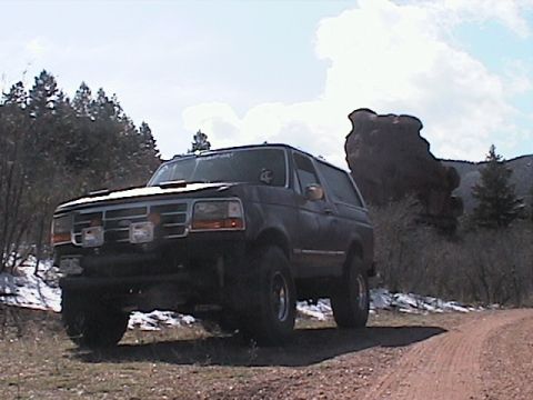1993 Ford Bronco - Dark SUV parked on a dirt road with trees and a large rock formation in the background.