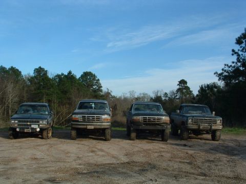 1993 Ford Bronco - Four trucks are lined up in a dirt lot with trees in the background.