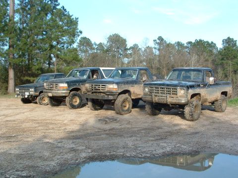 1993 Ford Bronco - Several lifted trucks, including a 1993 Ford Bronco, are parked in a muddy outdoor area.
