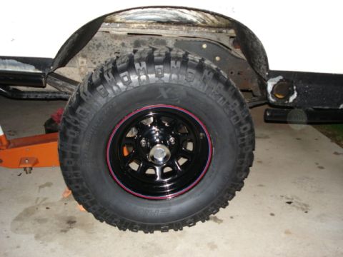 1987 Ford Bronco - Close-up of a vehicle's off-road tire and black wheel with red trim, showing part of the fender and frame.
