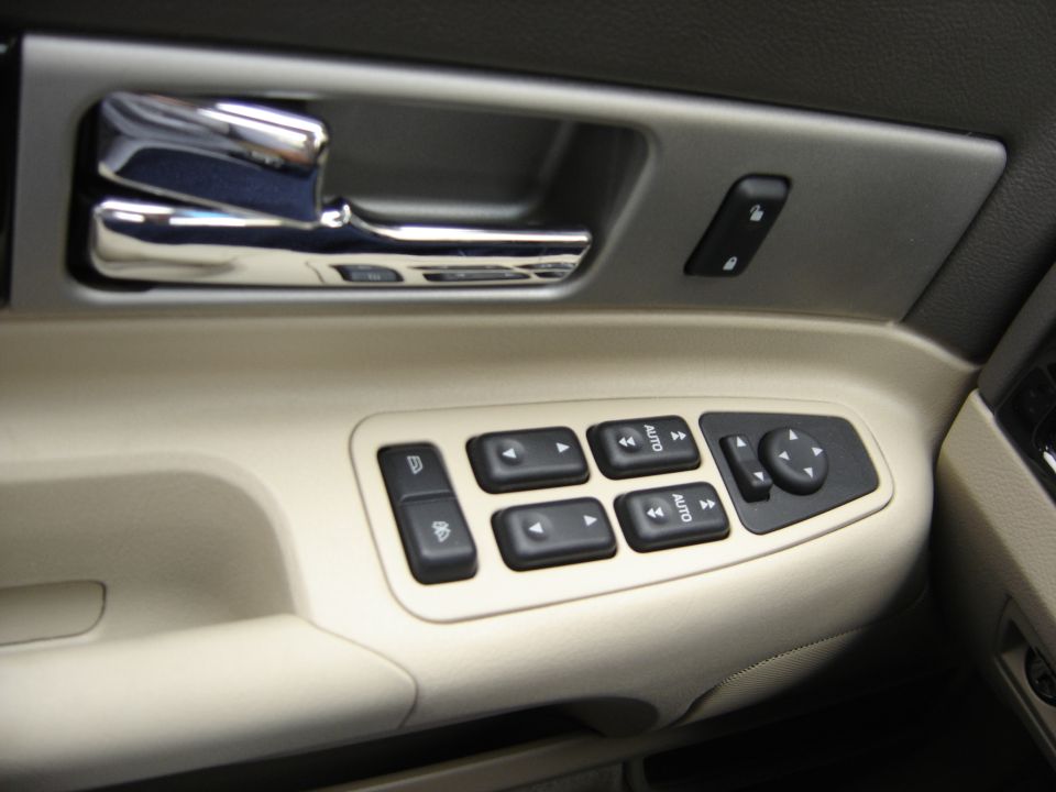 Car interior door panel with chrome handle, window controls, and mirror adjustment buttons. Beige and gray trim