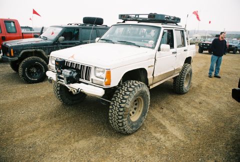 1995 Ford Bronco - A white Jeep Cherokee XJ is parked on a dirt lot with other vehicles and people around.