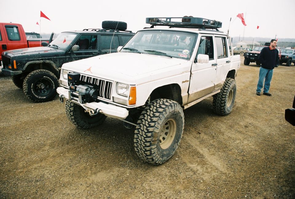 White Jeep Cherokee XJ with large tires and a winch, parked on dirt next to a black Jeep
