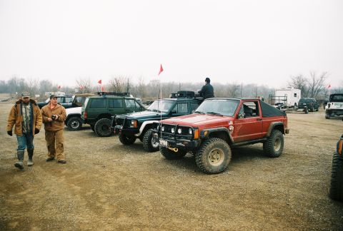 1995 Ford Bronco - A group of off-road vehicles, including a red Jeep Cherokee Chief, are parked in a lot.