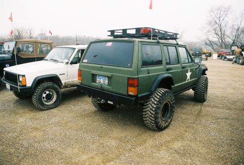 1995 Ford Bronco - Two Jeep Cherokee XJs are parked outdoors on a gravel lot, with other vehicles visible in the background.