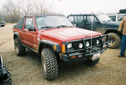 1995 Ford Bronco - A red Jeep Comanche pickup truck is parked outdoors with other vehicles.