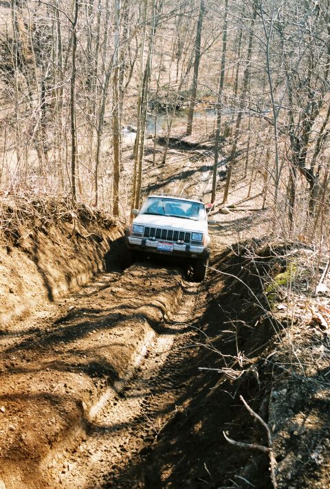 1995 Ford Bronco - A white Jeep Cherokee XJ navigates a challenging muddy trail through a wooded area.