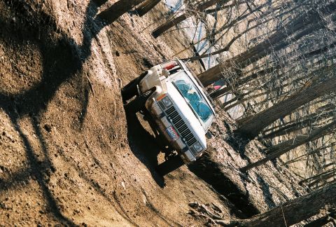 1995 Ford Bronco - A white Jeep Cherokee navigates a challenging muddy incline amidst trees.